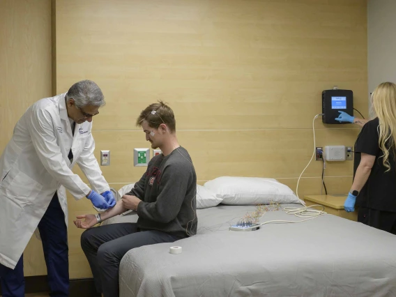 A doctor applies sensors to the arm of a person sitting on a bed in a sleep lab while another person monitors the sensor output on a small digital device on the wall.