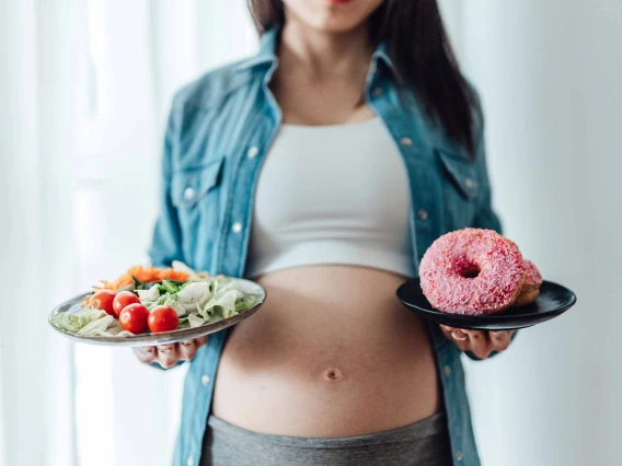 A pregnant woman stands with one plate of salad and another with a donut