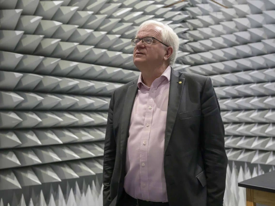 A man in a dress shirt and jacket stands in a small room with cone-shaped acoustic tiles on all the walls. 