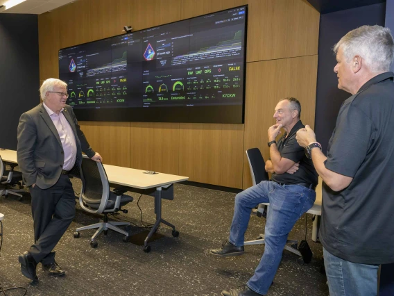 Three men stand in a room with two large monitors on the wall. 