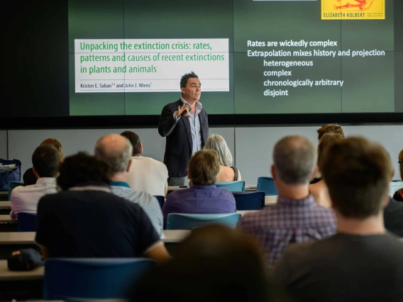 A man stands in front of an audience giving a presentation with a projection on the screen behind him.