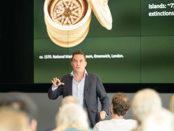 A man stands in front of an audience giving a presentation with a projection on the screen behind him. 