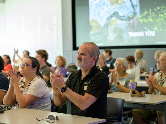 An audience sitting at rows of tables applauds. 