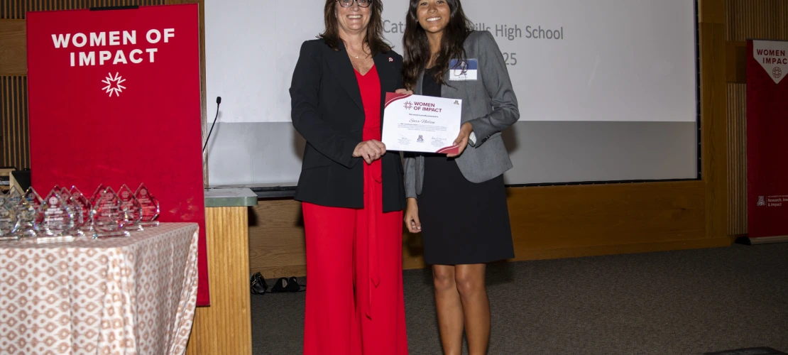 Two women holding a certificate pose for a photo in front of a projector screen.