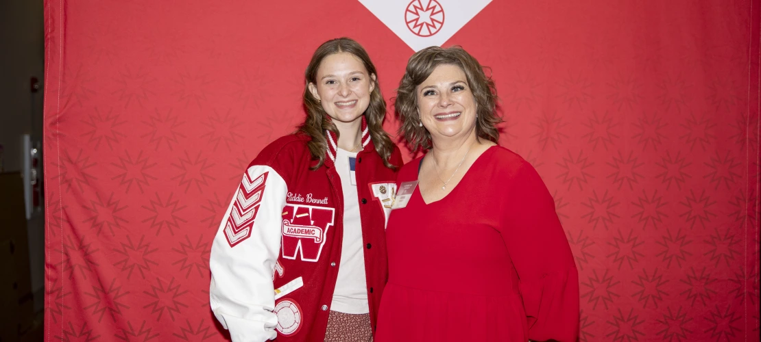 Two women pose for a photo in front of a red backdrop.