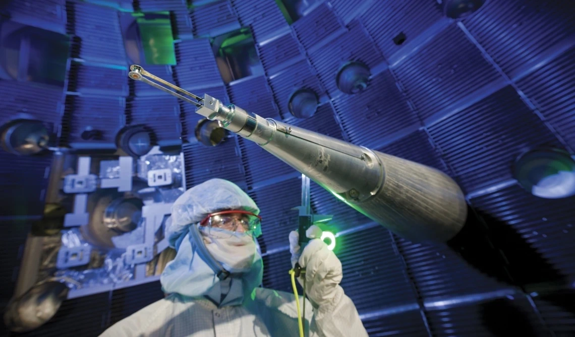 A technician in full cleanroom attire, including a face shield and hood, stands near a long, conical instrument used to position fuel targets inside the National Ignition Facility. The background shows the interior of the target chamber, with glowing blue lighting and metal structures creating a high-tech atmosphere.