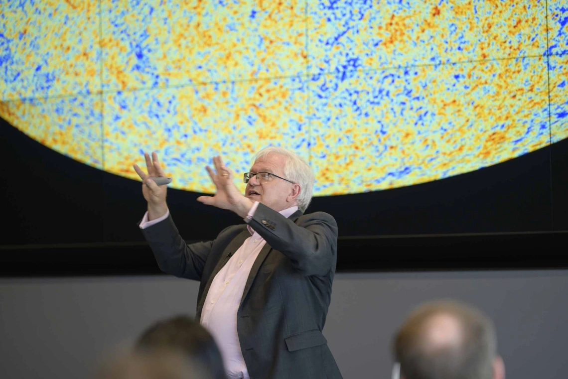 A man with white hair holds his hands in the air as he presents to an audience with a large circular projection of yellow, orange and blue dots behind him. 