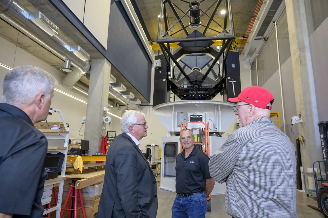 Four men stand while talking in a large room with a giant telescope in the background. 