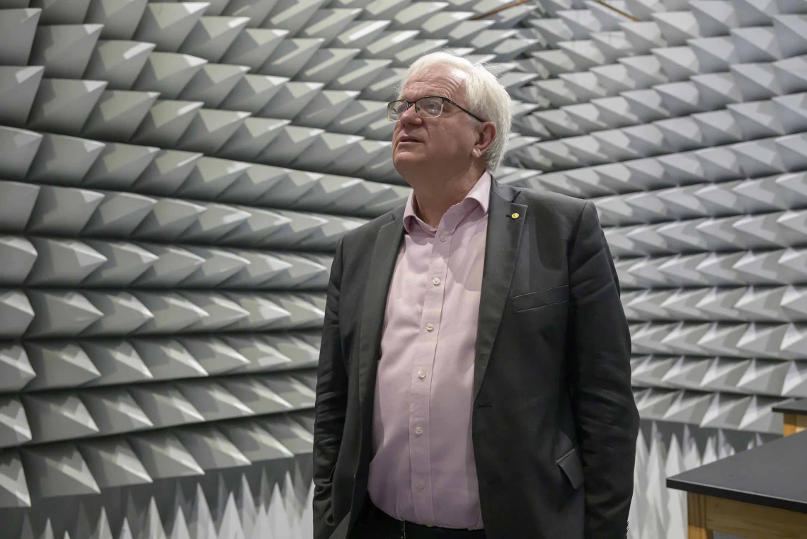 A man in a dress shirt and jacket stands in a small room with cone-shaped acoustic tiles on all the walls. 