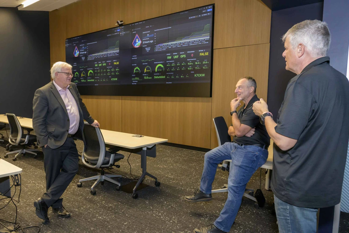 Three men stand in a room with two large monitors on the wall. 