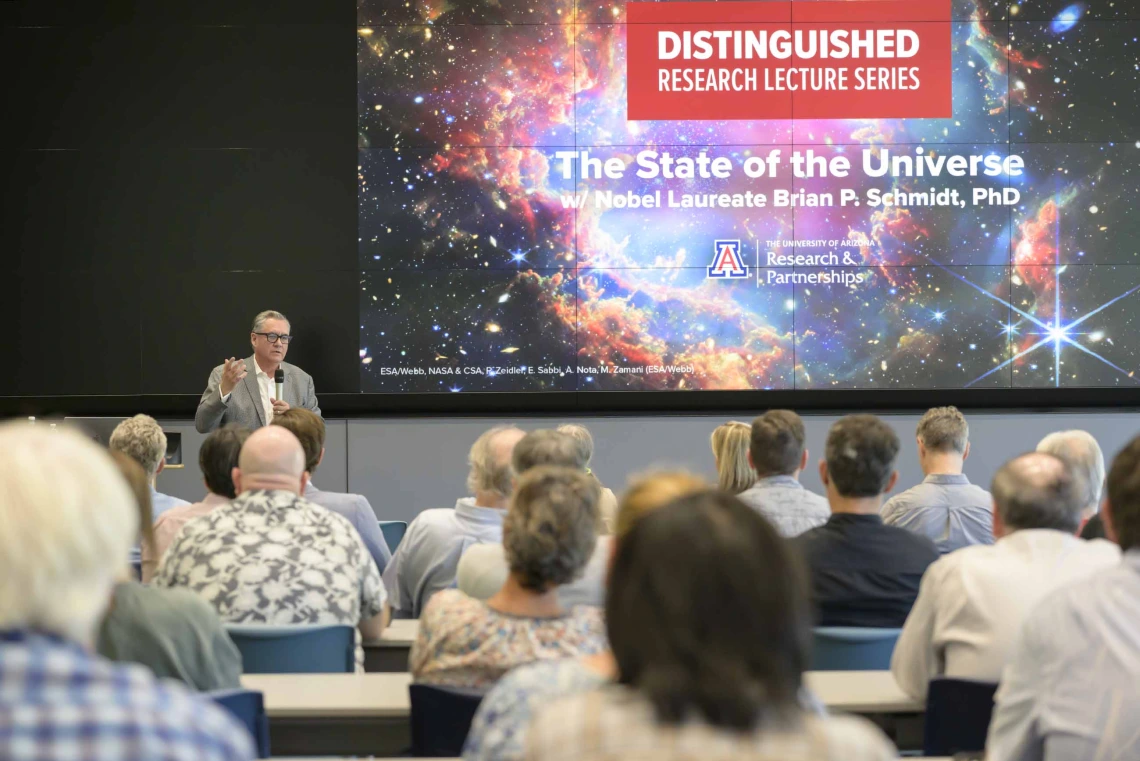 View from the back of a lecture hall over the heads of the audience with a projection of space with stars and text that reads, “Distinguished Research Lecture Series”. 