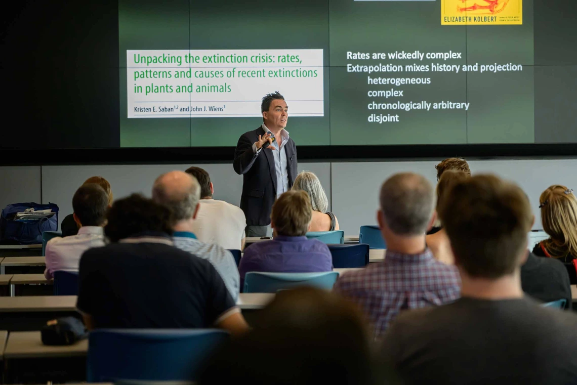 A man stands in front of an audience giving a presentation with a projection on the screen behind him.