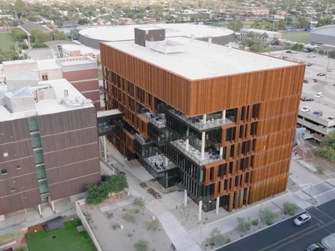 An aerial view of a copper-sided building with multiple levels of outdoor balconies. The building is attached to another via a walkway. Cars can be seen driving on the street outside.