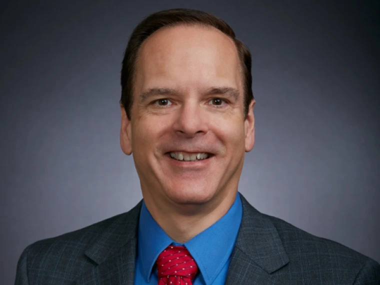 Smiling man in a blue shirt, red tie, and dark gray blazer posed against a dark gradient background, facing the camera in a professional headshot.